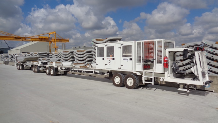Large white trailer with machinery on an industrial site, with stacked concrete segments and a yellow crane in the background.